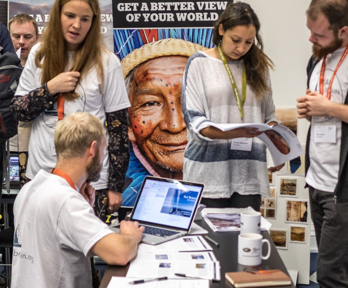 People stand and talk as one kneels to typr on a computer, surrounding a onfrence fair stand.