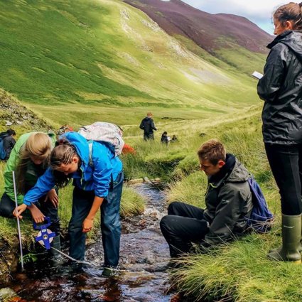 A group of students standing in a shallow upland stream, using a measuring tape to measure the width of the stream. In the background are green valley sides.