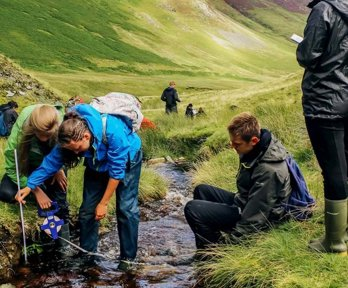 A group of students standing in a shallow upland stream, using a measuring tape to measure the width of the stream. In the background are green valley sides.