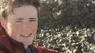 Headshot of Evan Chick. He is outdoors on a sunny day. In the background lays a large pile of rocks.