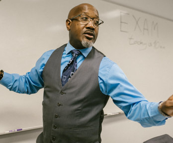 A teacher stands at the front of a classroom and speaks whilst writing on a whiteboard