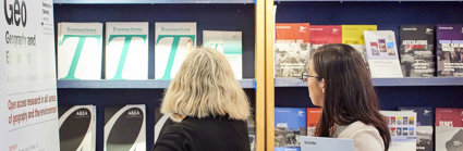 Two people standing in front of a bookcase filled with various Society journal publications such as Transactions, Geo and Area.