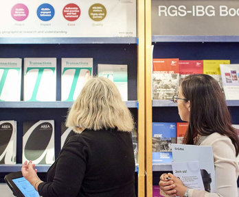 Two people standing in front of a bookcase filled with various Society journal publications such as Transactions, Geo and Area.