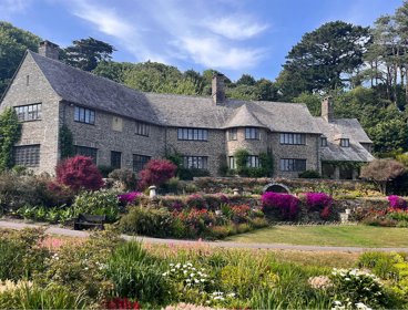 A large brick country house in the middle of a forest and a garden full of flowers in bloom.