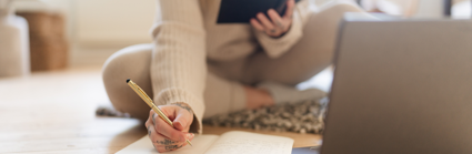 A person sits on a wooden reading a book being held in one hand and writing in a note book with their other hand. A silver laptop is laying on the ground on front of them