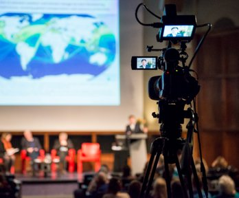 A panel of speakers on stage viewed from the back of a theatre, with a camera filming the event in the foreground.