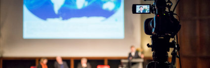A panel of speakers on stage viewed from the back of a theatre, with a camera filming the event in the foreground.