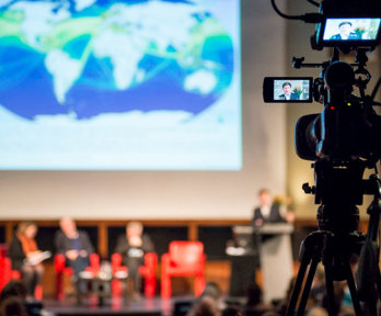 A panel of speakers on stage viewed from the back of a theatre, with a camera filming the event in the foreground.