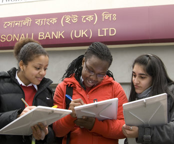 A group of three female students with clipboards, collecting information in a high-street area. They are standing outside a bank.