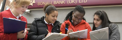 A group of three students with clipboards, collecting information in a high-street area. They are standing outside a bank.