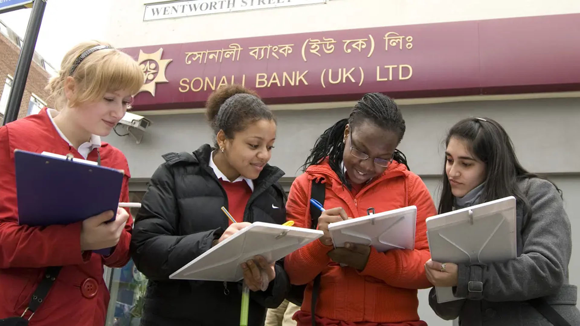 A group of three students with clipboards, collecting information in a high-street area. They are standing outside a bank.