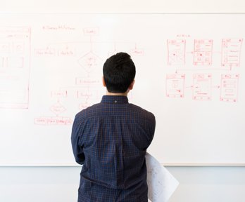 Man holding a piece of paper looking at a whiteboard with diagrams drawn on it.