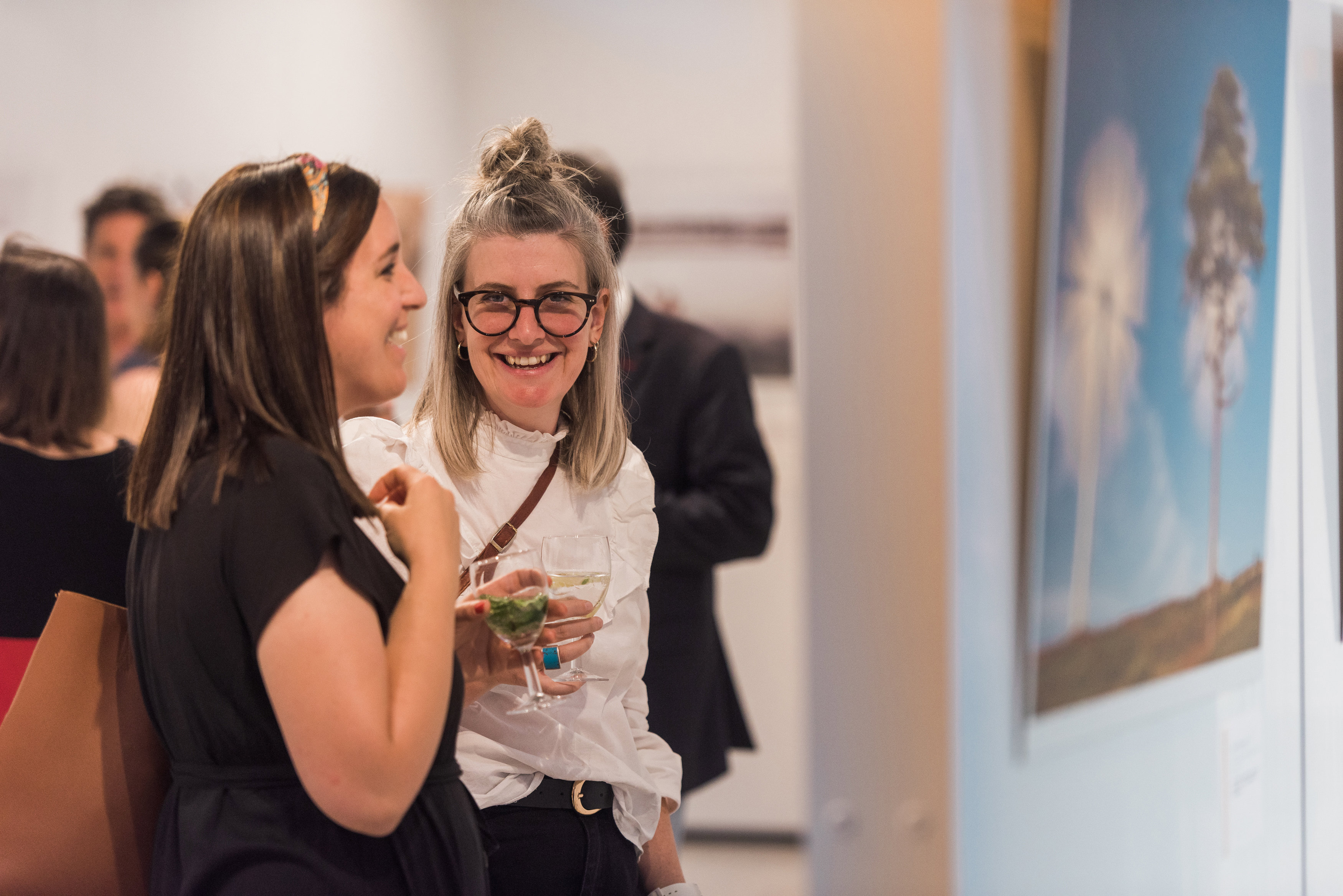 Two young people holding a drink while at a photograph exhibition. One person smiles directly at the camera.