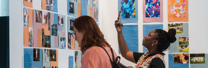 Two people stand in front of a board with posters. One is reading a caption while the other takes a photo with a smartphone.