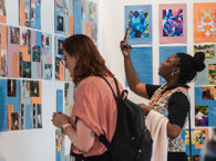 Two people stand in front of a board with posters. One is reading a caption while the other takes a photo with a smartphone.