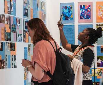 Two people stand in front of a board with posters. One is reading a caption while the other takes a photo with a smartphone.