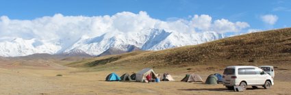 A group of small tents and a white van in a wide unvegetated landscape in front of snow covered mountains.