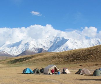 A group of small tents and a white van in a wide unvegetated landscape in front of snow covered mountains.