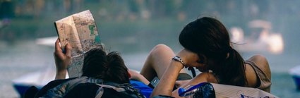 Two people lay down on the grass looking at a map whilst they both rest their heads on their backpacks