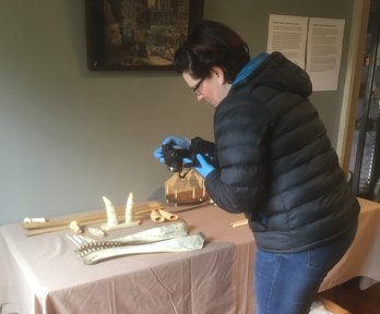 Artefacts laid out on a cloth covered table. A person wearing gloves is stood in front of the table holding a camera to take photos of the artefacts.