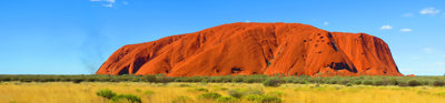 Bright colours define Uluru, with orange soil in the foreground scattered with green grass clumps, and a bright blue sky with a few small clouds. Uluru stand in the distance and is bright orange in colour.