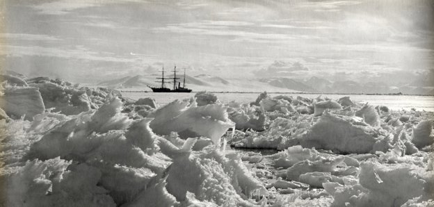 Black and white photo taken in Antarctica of ice in the foreground and a large vessel on the water in the background