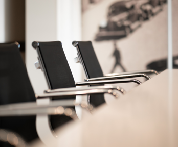 A row of boardroom chairs are neatly pushed under one side of a table. The larger boardroom backdrop is blurred.