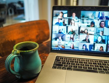 A laptop open wiht an online meeting showing beside coffee cup