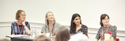 Four speakers sitting at a table in front of an audience, looking over to the right to a person not pictured.