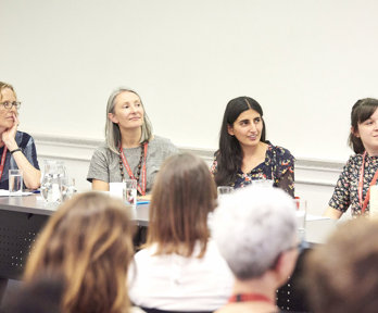 Four speakers sitting at a table in front of an audience, looking over to the right to a person not pictured.