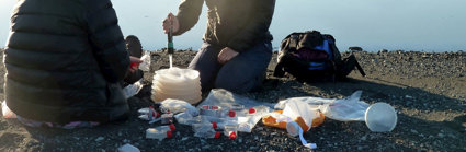 Two people sat beside a glacial lake taking measurements. They are surrounded by small transparent tubes and other measuring equipment.