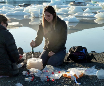 Two people sat beside a glacial lake taking measurements. They are surrounded by small transparent tubes and other measuring equipment.