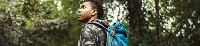 Person with a backpack on in a forest writing something down in a notebook while looking up. 