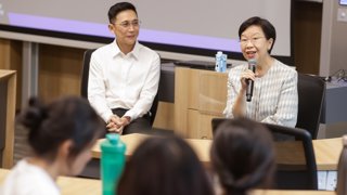 A female professor who is holding a microphone and addressing a group of university staff and students.