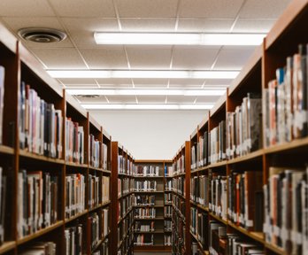 Two rows parallel libary shelves with a perpendicular row the can be seen between the two. All shelves are stacked full of books.