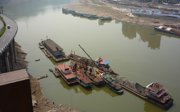 A brownish river with several industrial boats in it, with a highway on the left side and empty plots of land on the right.