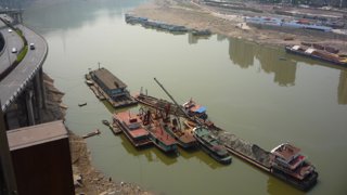 A brownish river with several industrial boats in it, with a highway on the left side and empty plots of land on the right.