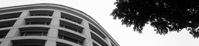 A modern building seen from below. In the top corner are some tree branches. The photo is in black and white.