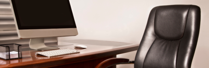 A leather office chair with varnished brown wood arm rests sit empty on front of a brown wooden desk. A laptop, keyboard, mouse  and a stack of note paper sit on the desk.