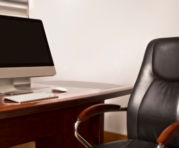 A leather office chair with varnished brown wood arm rests sit empty on front of a brown wooden desk. A laptop, keyboard, mouse  and a stack of note paper sit on the desk.