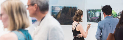 People viewing a photographic exhibition displayed on white panels in a modern pavilion. The photos show forests and trees.