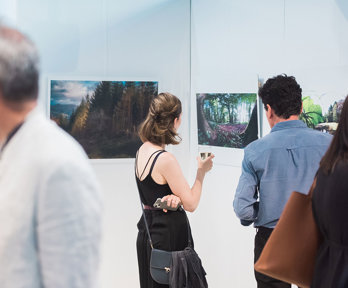 People viewing a photographic exhibition displayed on white panels in a modern pavilion. The photos show forests and trees.