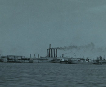 Black and white photograph of a river in the foreground and a factory in the background.