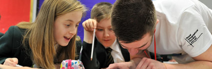 Two young children sit at their desk in a classroom looking at a small map on their desk with an adult pointing at something on the map.