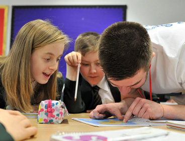 Two young children sit at their desk in a classroom looking at a small map on their desk with an adult pointing at something on the map.