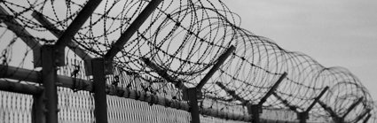 View of the top of a barbed wired fence in front of a dark cloudy sky. The image is in black and white. 