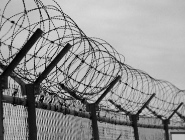 View of the top of a barbed wired fence in front of a dark cloudy sky. The image is in black and white. 