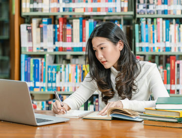 Student in a library looking at a laptop screen while writing notes with in the background various books on a book shelf.