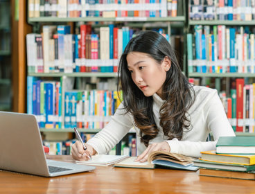 Student in a library looking at a laptop screen while writing notes with in the background various books on a book shelf.