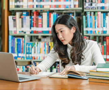 Student in a library looking at a laptop screen while writing notes with in the background various books on a book shelf.